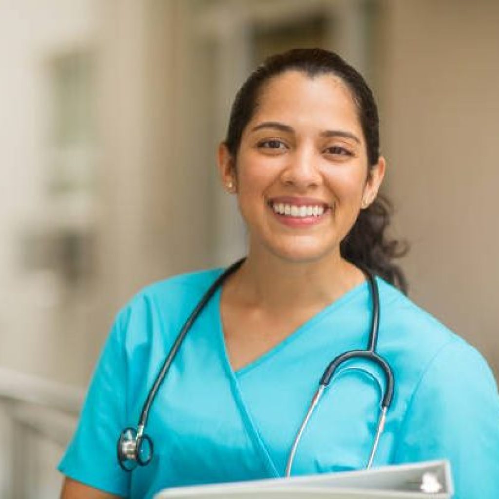 Smiling female healthcare professional looks at the camera while in hospital hallway. She is standing with her arms crossed.
